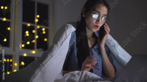 Young Asian woman working in the bed use smartphone talking team and working with a laptop late at night