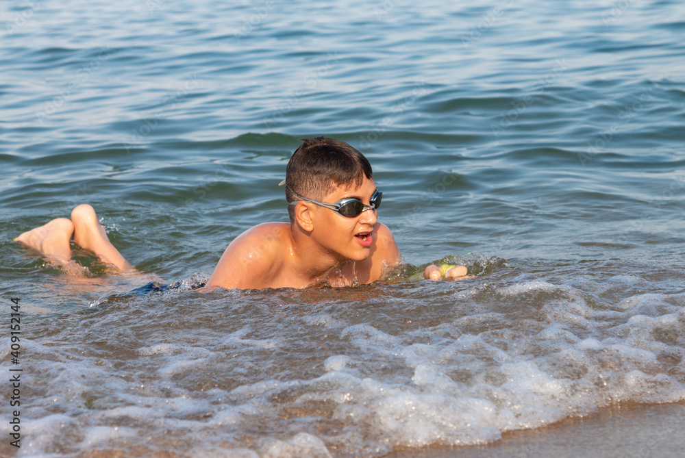 Teenager in goggles swims in the sea with big splashes. Cute boy enjoys swimming in the ocean.  Holidays, vacation with kids