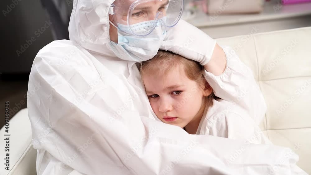 doctor in protective suit, mask, gloves and glasses calms down little sick girl.