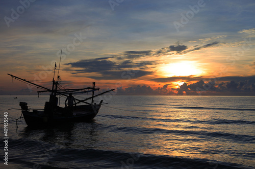 The silhouette of a fishing boat floats on the water in the morning, the sun is rising, the sky is beautiful blue and orange.