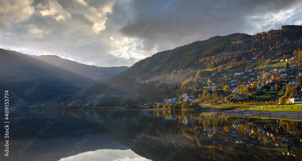 A panoramic view of the lake in Voss Norway, the water is still mirrored and the sun shines down in the evening, the house on the hill with beautiful trees in autumn.