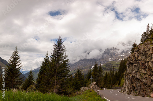 A panoramic spring view of Swiss Alps at high elevation. Clouds move by the summits of steep cliffs and mountains. Pine trees, grass and flowers are seen alongside a curvy mountain road.