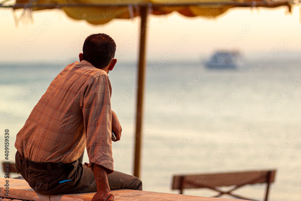 A Jordanian man wearing long sleeve shirt is sitting on a wall by the beach in Aqaba, Jordan. He is watching the ships pass by the gulf of Aqaba, red sea with his arm on his knee. Peaceful scene.