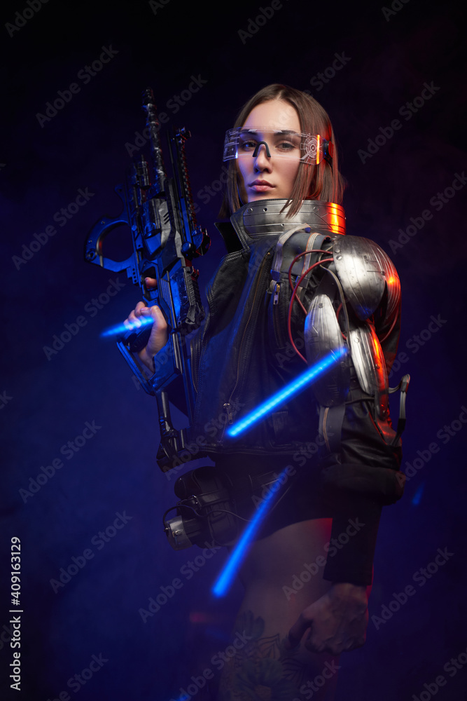 Naklejka premium Tattoed martial woman in cyberpunk style dressed in stylish black clothing. Fashionable and sexy female soldier holding a futuristic rifle and posing in dark background with blue lights.
