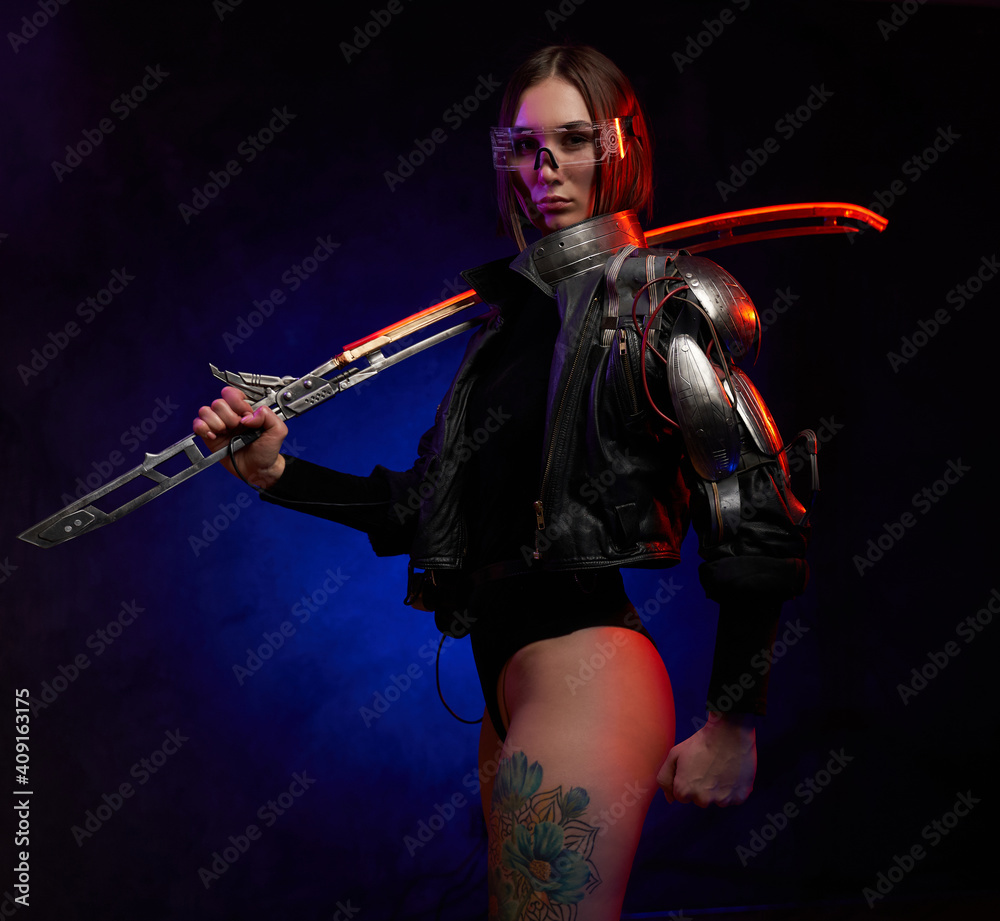 Fashionable woman killer with short haircut holding futuristic sword on her shoulder. Martial woman in black costume with eyewear posing in dark background with lights.