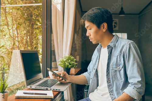 Left View Young Asian Casual Businessman in Denim or Jeans Shirt Using Smartphone and Laptop in Coffee Shop. Casual Businessman Working with Technology in Vintage Tone