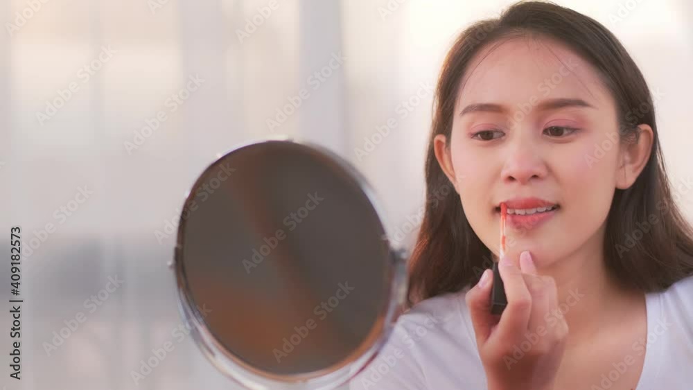 Beautiful attractive young Asian woman happy smiling doing beauty make up tutorial putting lipstick on lips looking at mirror and camera cheerful energetic at home living lifestyle with sunset light Beautiful attractive young Asian woman happy smiling doing beauty make up tutorial putting lipstick on lips looking at mirror and camera cheerful energetic at home living lifestyle with sunset light