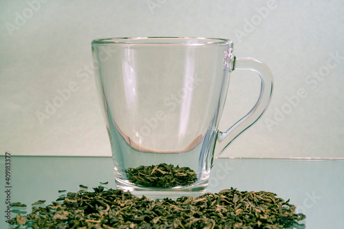 A cup with green tea leaves is on the table. Dry green tea leaves are scattered nearby. Green tea. Transparent cup. Copy space.
