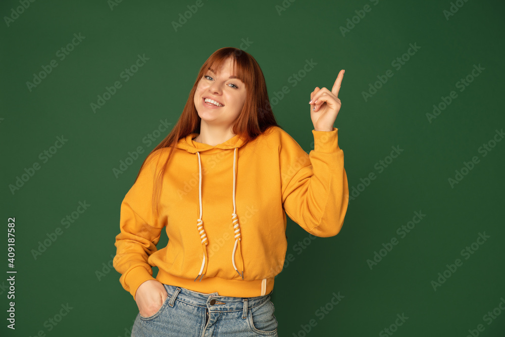 Pointing. Caucasian woman's portrait isolated on green studio background with copyspace. Beautiful female model in yellow sweatshirt. Concept of human emotions, facial expression, sales, ad, fashion. Pointing. Caucasian woman's portrait isolated on green studio background with copyspace. Beautiful female model in yellow sweatshirt. Concept of human emotions, facial expression, sales, ad, fashion.