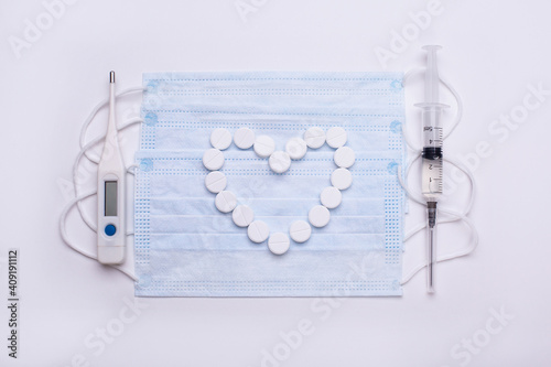 A syringe with a vaccine and a thermometer lie on the medical masks, and in the middle of the masks of tablets is laid out a heart. Vaccination against coronavirus.