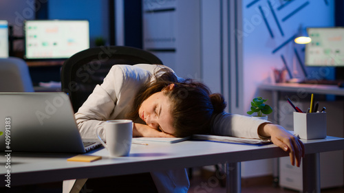 Exhausted overload business woman falling asleep on desk with open laptop monitor while working in start up company office. Employee using modern technology network wireless doing overtime sleeping photo