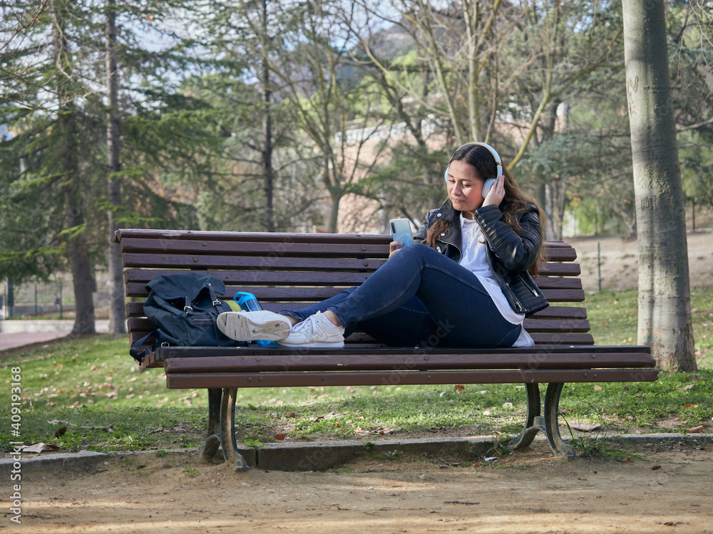 Young female with headphones using her smartphone while sitting on a wooden bench in the park Young female with headphones using her smartphone while sitting on a wooden bench in the park