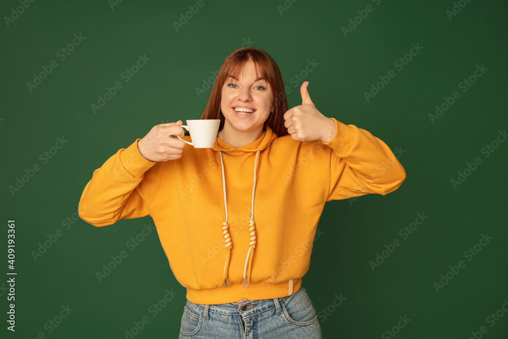 Enjoying coffee, tea. Caucasian woman's portrait on green studio background with copyspace. Beautiful female model in yellow sweatshirt. Concept of human emotions, facial expression, sales, ad