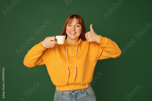 Enjoying coffee, tea. Caucasian woman's portrait on green studio background with copyspace. Beautiful female model in yellow sweatshirt. Concept of human emotions, facial expression, sales, ad © master1305