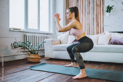 Young fit woman in sportswear do squat exercise indoors in living room. Online indoors workout. Training at home photo