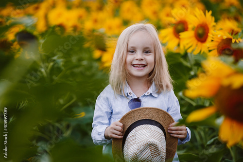 Little girl of six years old poses for the camera in a field of sunflowers, laughs and holds a hat in her hands