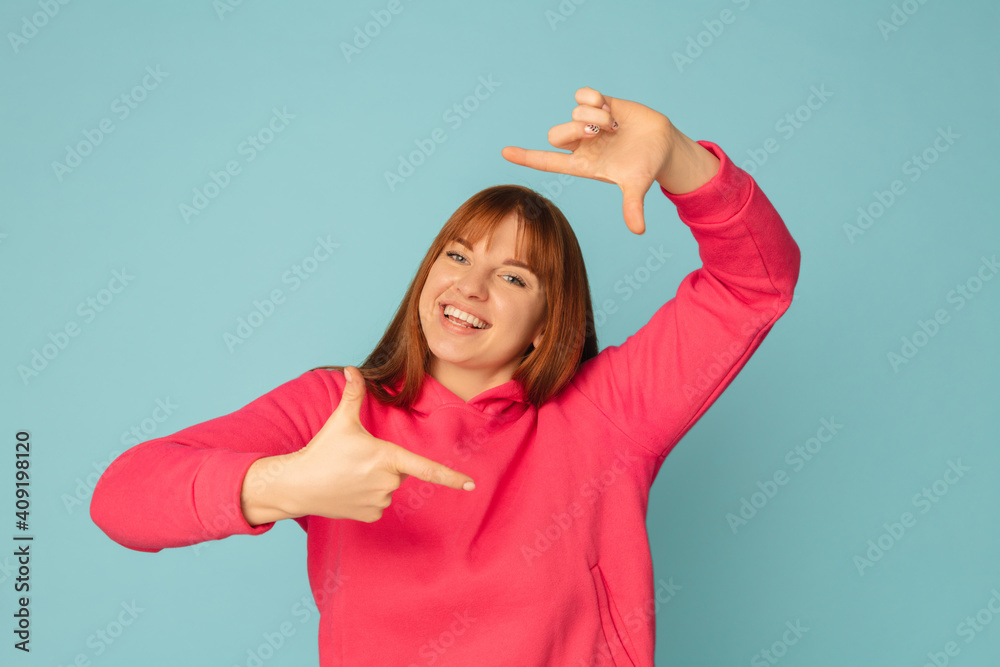 Fototapeta premium Framing. Caucasian woman's portrait isolated on blue studio background with copyspace. Beautiful female model in pink sweatshirt. Concept of human emotions, facial expression, sales, ad, fashion.
