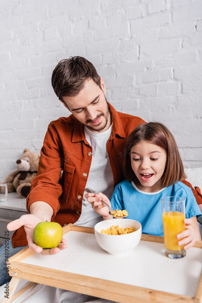 excited girl eating cornflakes near father holding apple in hospital excited girl eating cornflakes near father holding apple in hospital