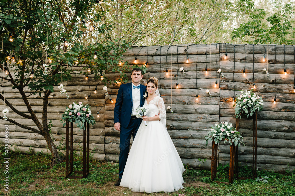 Wedding couple in magical evening forest decorated light garlands. Ceremony with lanterns and lamps decorated flowers. Bride and groom on background of bulb lights.