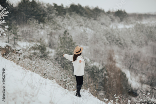 Happy girl traveler with hands up standing on the top of mountain and looking on beautiful winter snowy landscape. 