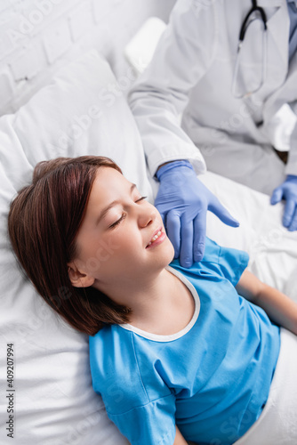 doctor touching child lying in hospital bed with closed eyes, blurred background