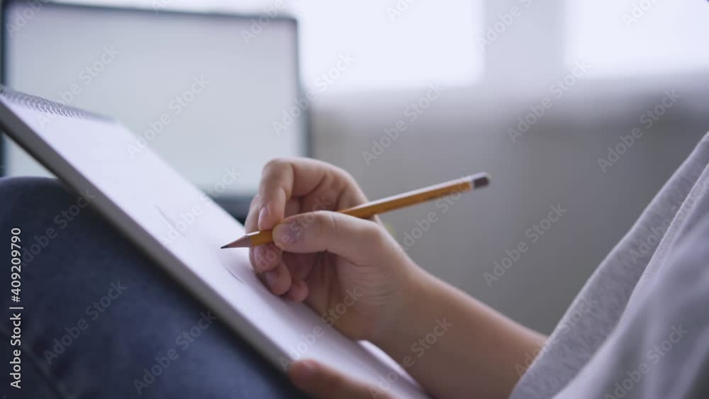 Creative girl artist draws in sketchbook with pencil and turns sheets of paper sitting at home against blurred windows with bright light close view Creative girl artist draws in sketchbook with pencil and turns sheets of paper sitting at home against blurred windows with bright light close view
