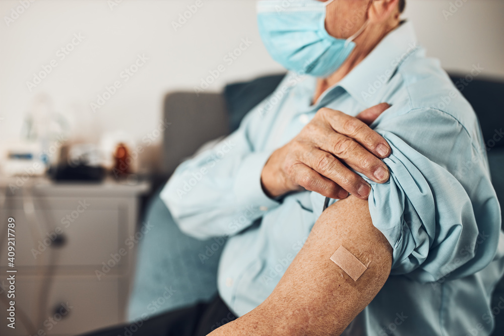 Senior man patient holding shirt sleeve up with a plaster in place of injection of vaccine. Covid-19 or coronavirus vaccination. Man wearing face mask Senior man patient holding shirt sleeve up with a plaster in place of injection of vaccine. Covid-19 or coronavirus vaccination. Man wearing face mask