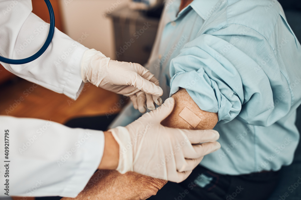 Doctor putting a plaster in place of injection of vaccine to senior man patient. Covid-19 or coronavirus vaccination. Physician wearing white coat and gloves using face mask Doctor putting a plaster in place of injection of vaccine to senior man patient. Covid-19 or coronavirus vaccination. Physician wearing white coat and gloves using face mask