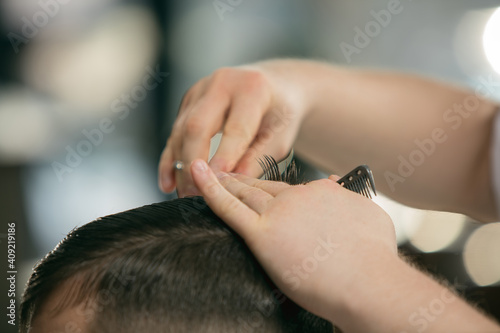 Close up hands of master barber, stylist with sccissors does the hairstyle to guy, young man. Professional occupation, male beauty concept. Cares of hair of client. Soft colors and focus, vintage.