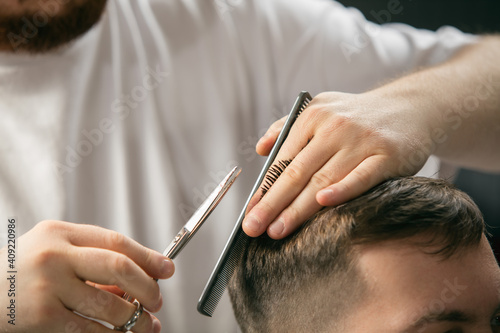 Close up hands of master barber, stylist with sccissors does the hairstyle to guy, young man. Professional occupation, male beauty concept. Cares of hair of client. Soft colors and focus, vintage. photo