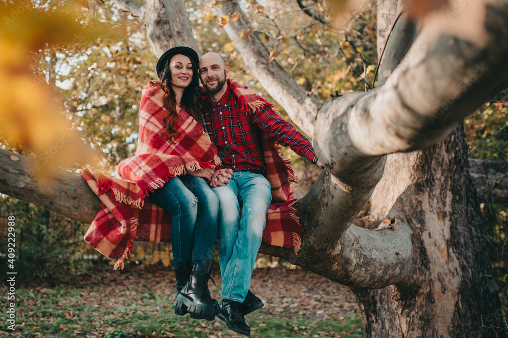 A man and a woman are sitting on a tree branch, wrapped in a blanket and looking forward