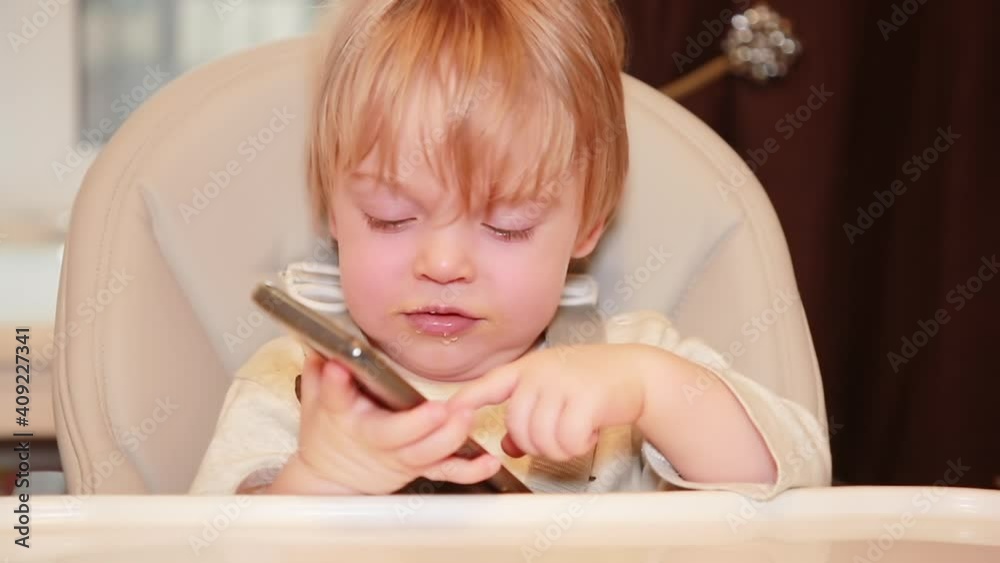 Portrait of a child in a chair with a smartphone. The kid learns to use the phone. Children and new technologies. Selective focus Portrait of a child in a chair with a smartphone. The kid learns to use the phone. Children and new technologies. Selective focus