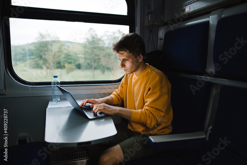 A freelancer man sitting in a train at a table and working on a laptop with a serious face. Freelancer uses laptop on the way by train. Working in travel. © bodnarphoto