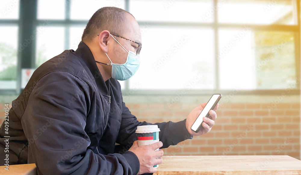 Side view young businessman wearing a mask Wear a black sweater Sit at the table in the coffee shop and take your smartphone and a cup of coffee. Side view young businessman wearing a mask Wear a black sweater Sit at the table in the coffee shop and take your smartphone and a cup of coffee.