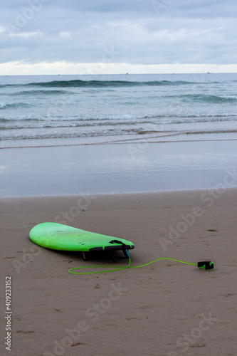 View of green surfboard on the shore of the beach, with the sea in the background, vertical, a cloudy day, in Cantabria, Spain