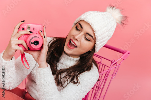 Fascinated brunette in knitted hat makes photo on camera instax. Portrait of young girl in pink trolley on isolated background photo