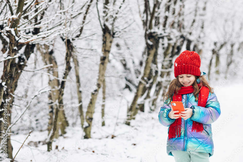 Little girl rolling up a funny video in smartphone. Happy child playing at winter forest use a smartphone look at the screen of a cell phone, watch cartoons