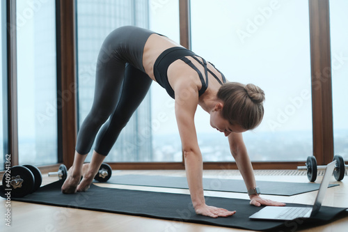 Portrait of a flexible and graceful female person in sportive clothing doing yoga on mat using a laptop in apartment.