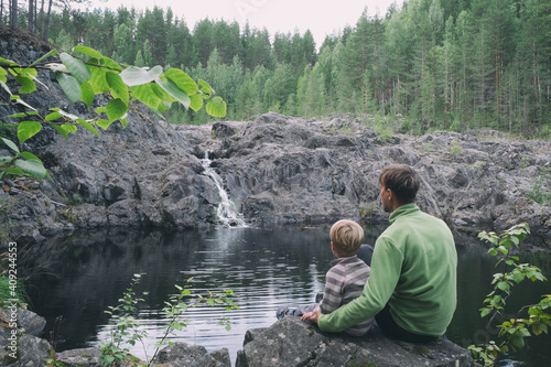 Father and son hikers travellers sitting near mountain waterfall. Peaceful and calmness scenic view. Active family outdoors, mindfulness living.