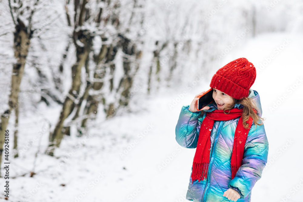 Pretty little child girl with curly hair in winter clothes talking on mobile phone smiling standing on the winter park near Christmas trees. Little child girl having conversation on her mobile phone.