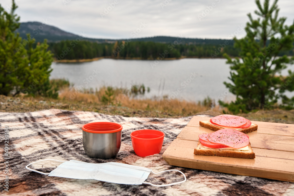 Sandwiches on the wooden stand with orange mugs from thermos with mask on the plaid. Cozy picnic on fresh air. Mountains are visible in the distance. Sandwiches on the wooden stand with orange mugs from thermos with mask on the plaid. Cozy picnic on fresh air. Mountains are visible in the distance.