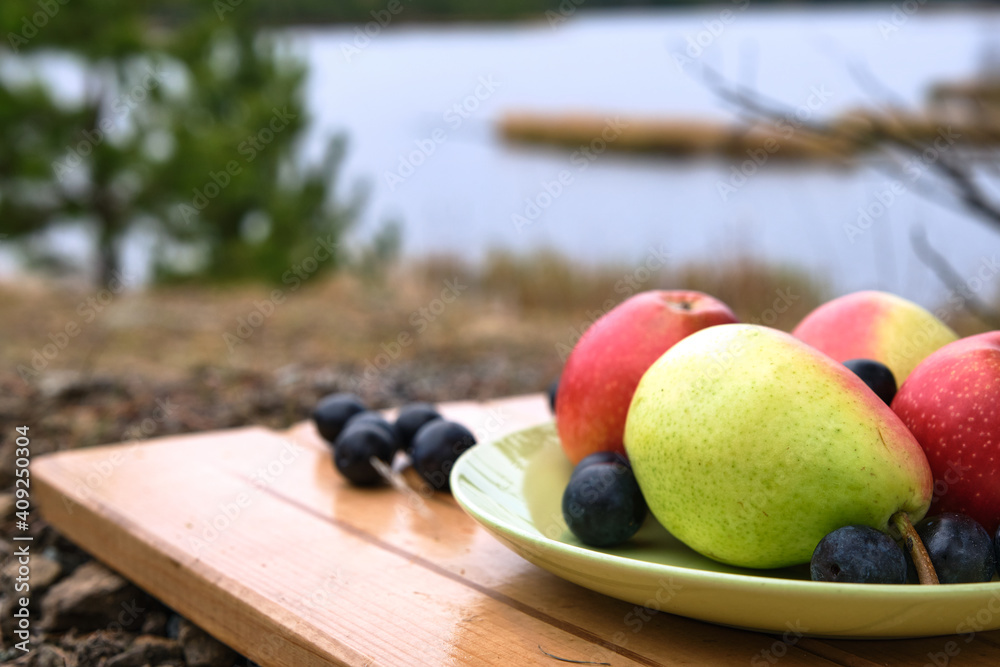 Apples, pears, grape lies on a green plate on a wooden stand. Cozy picnic on fresh air. Stone ground. Mountains are visible in the distance. Vegetarian healthy meal