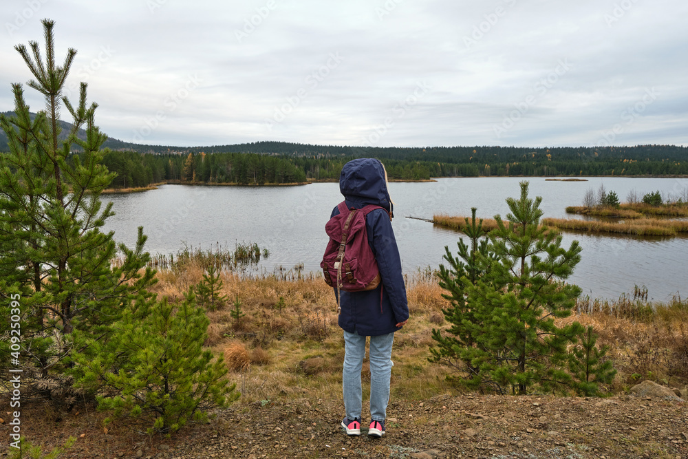 woman with backpack is standing in front of a river, forest and little island. little spruce grow near her. She put on a hoodie jacket. Solo female tourism. Lifestyle moment. woman with backpack is standing in front of a river, forest and little island. little spruce grow near her. She put on a hoodie jacket. Solo female tourism. Lifestyle moment.