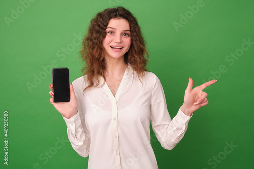 A girl in white clothes on a green background. The girl holds a switched-off phone in her hand, looks at the camera with a smile and points to an empty area with her index finger