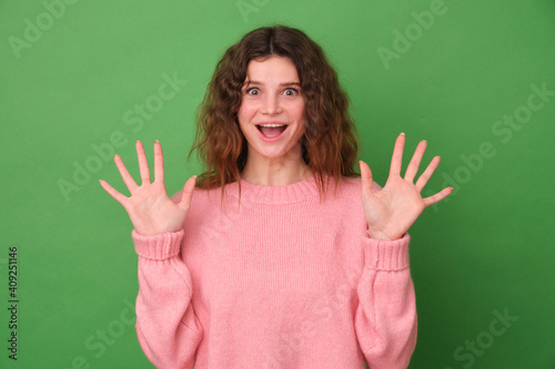 Overjoyed woman has curly Afro hair feels very happy raises her hands reacts to an amazing surprise wears a pink sweater poses on a green Studio background.