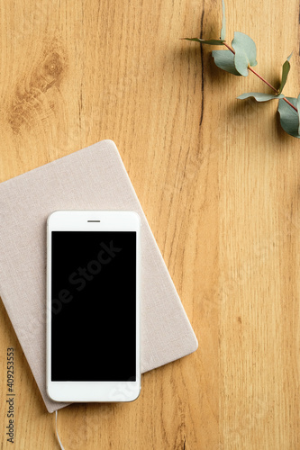 Mobile phone on wood table with paper notebook and eucalyptus leaf. Flat lay, top view home office desk.