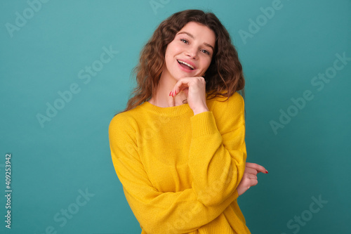 The cheerful beauty laughs sincerely, she has a cheerful expression on her face, has an interesting idea, holds her hand on a hoodie, enjoys life, poses indoors on a blue background of the studio