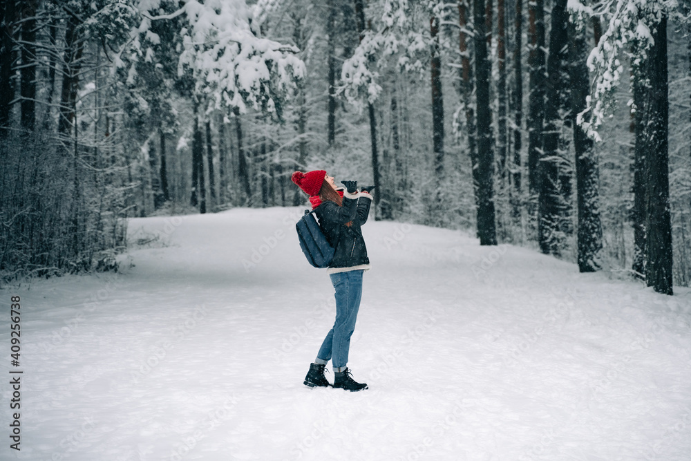 A beautiful woman in a red hat in a winter snowy forest is surprised, raises her hands up in the hope of catching a snowflake.