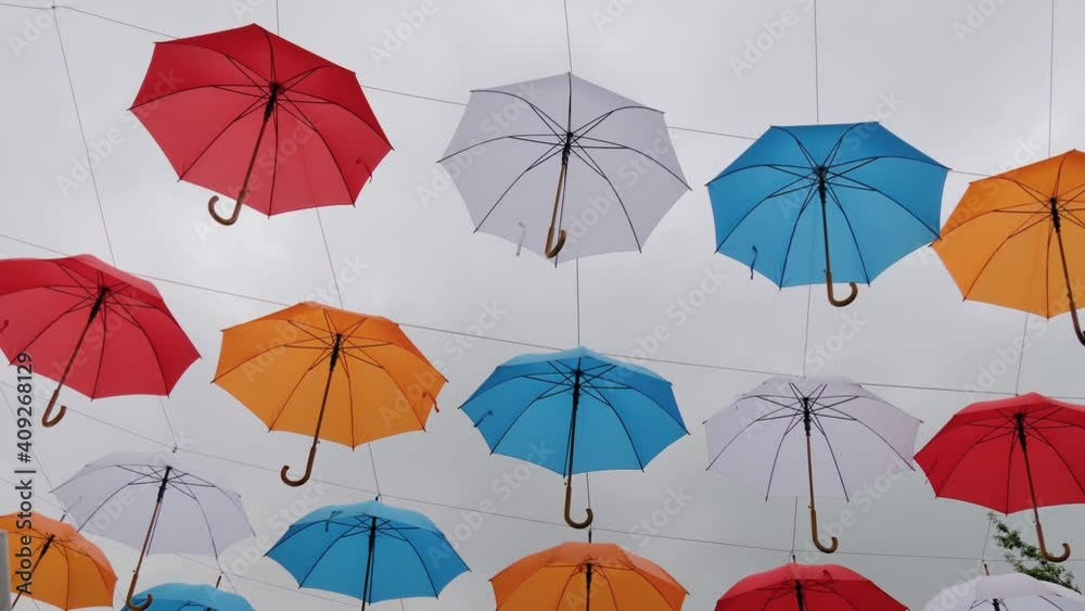 Slow motion: colorful umbrellas hanging against gray overcast sky and swaying in wind at summer city festival - low angle view. Street decoration, celebration, art, holiday concept