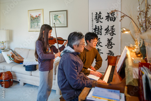 A father and uncle sit at piano while girl plays violin behind them photo